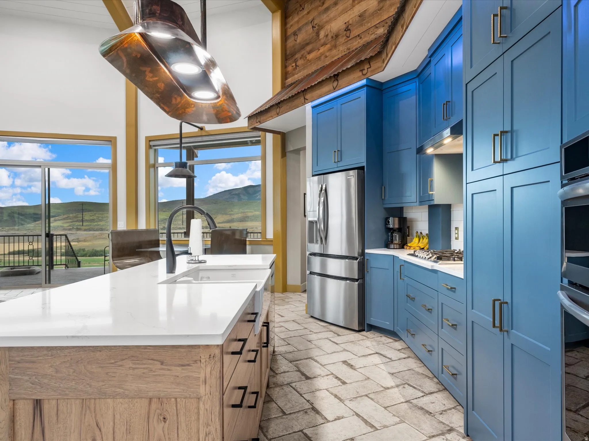 Contemporary kitchen featuring blue cabinetry, stainless steel appliances, a large center island with a white countertop and undermount sink, herringbone tile flooring, and oversized windows with sliding glass doors