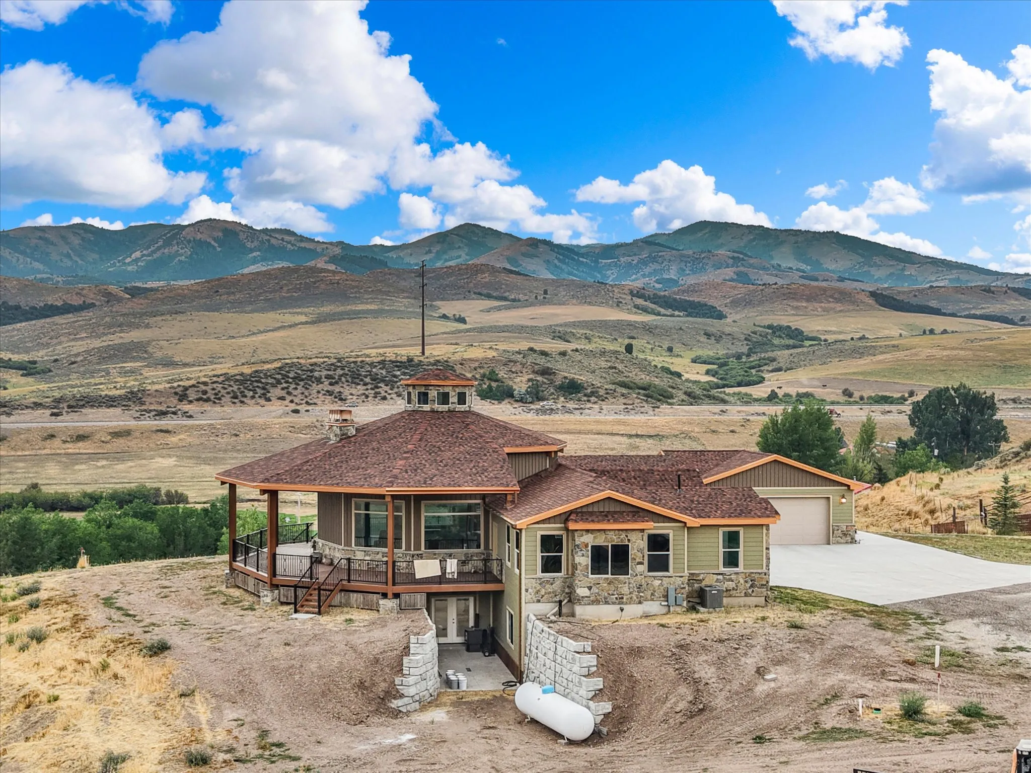 Mountain-side residence featuring a circular turret with multiple windows, extensive decking with glass panel railings, and a lower-level concrete patio