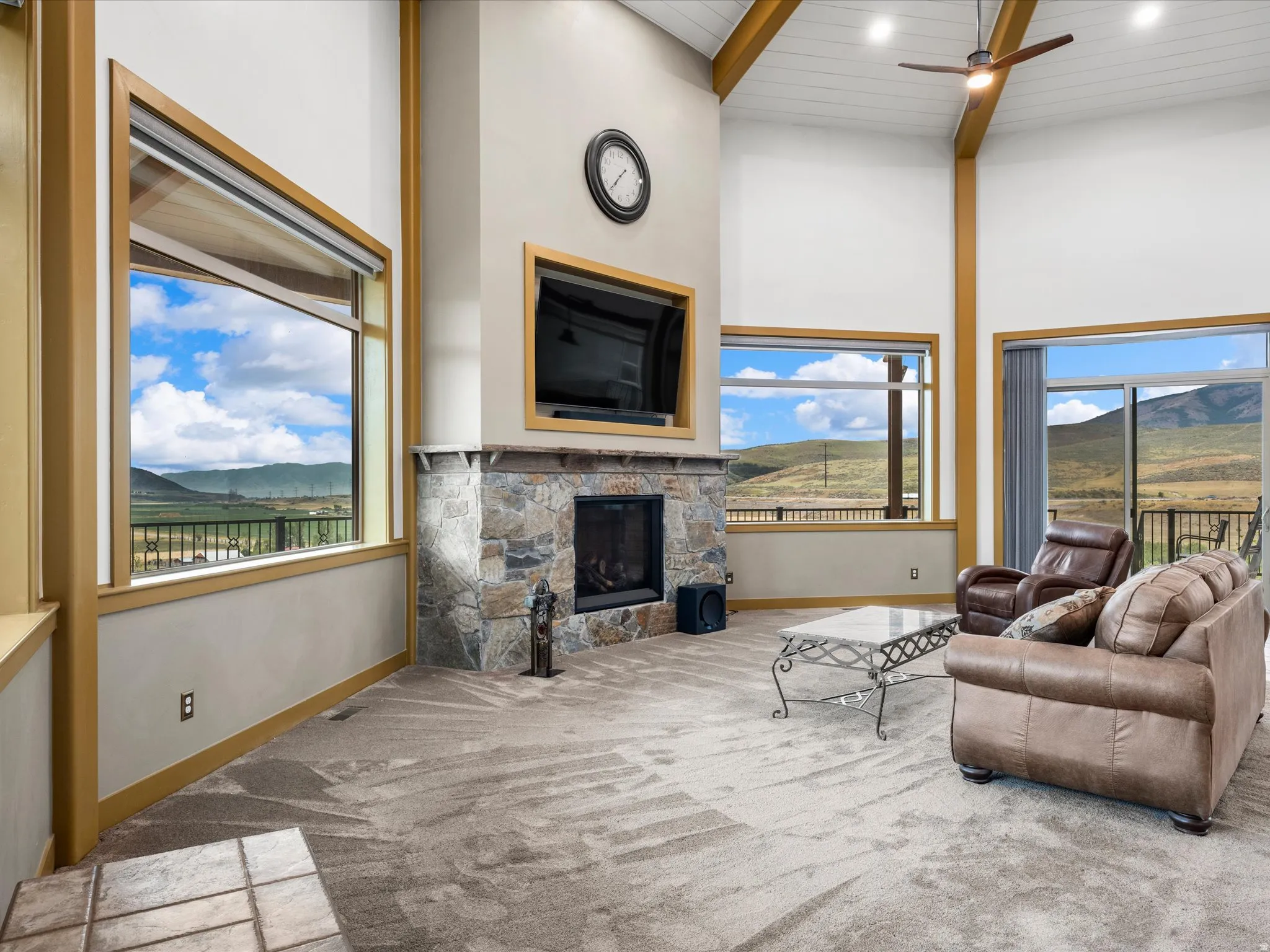 Vaulted ceiling living area featuring a stone-clad fireplace with mantel, expansive windows, a sliding glass door to an outdoor deck, exposed wood beams, and a ceiling fan