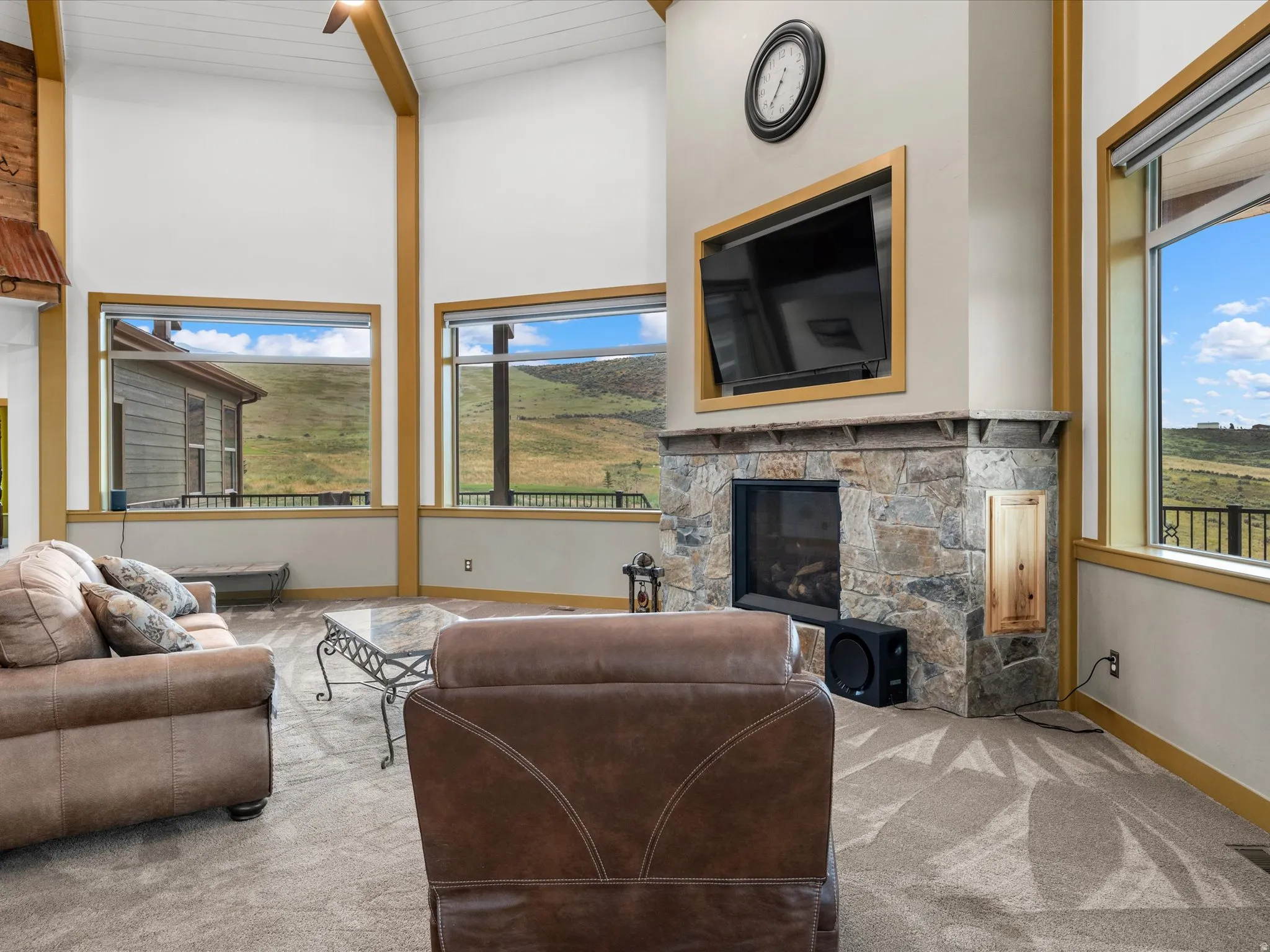 Vaulted ceiling with exposed wood beams, stone-clad fireplace, large picture windows, and light-toned carpeting