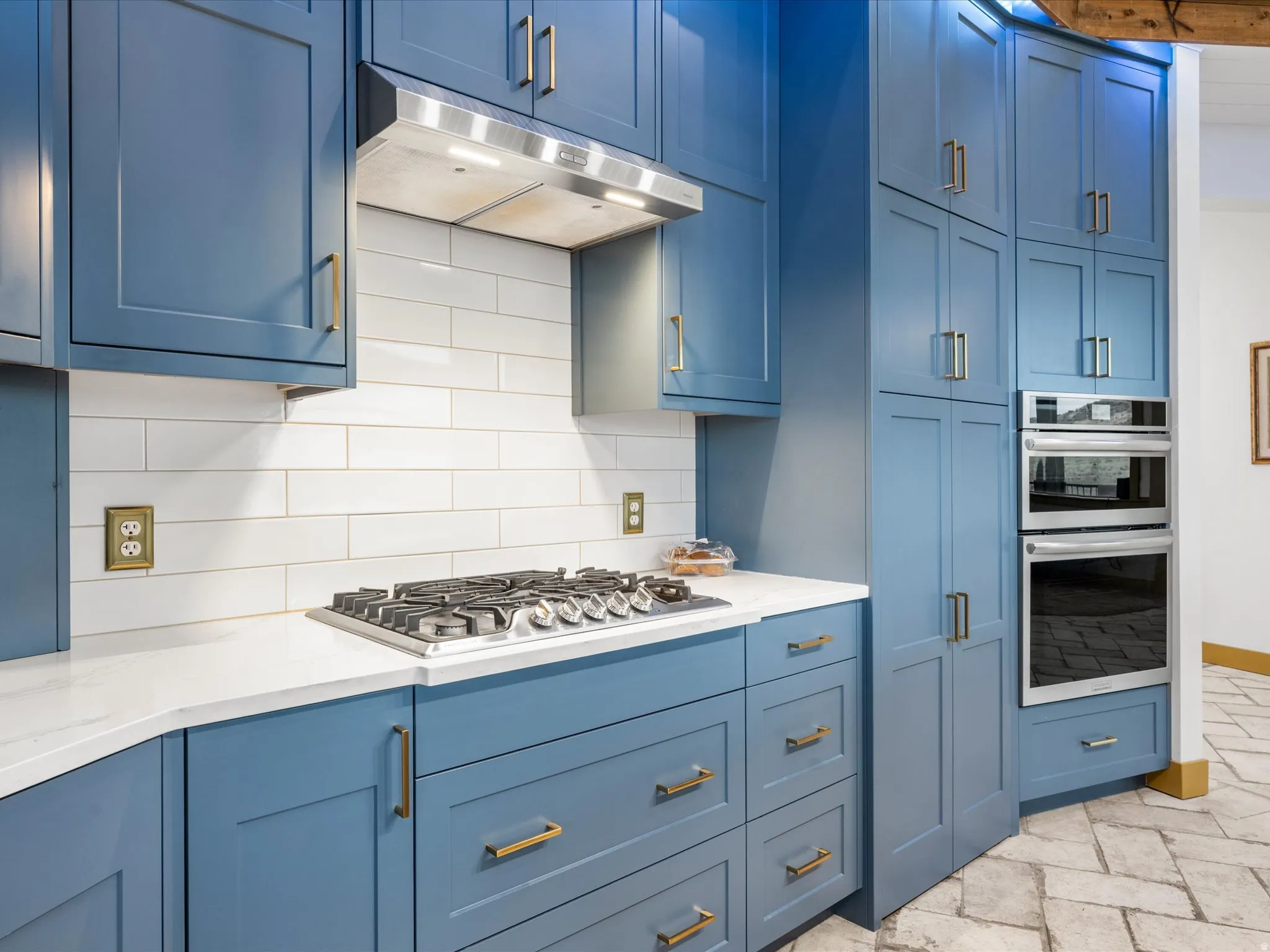 Kitchen featuring blue cabinetry with brass-tone hardware, stainless steel gas cooktop, white subway tile backsplash, white countertops, and double wall ovens