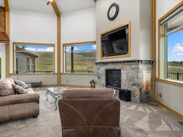 Vaulted ceiling with exposed wood beams, stone-clad fireplace, large picture windows, and light-toned carpeting