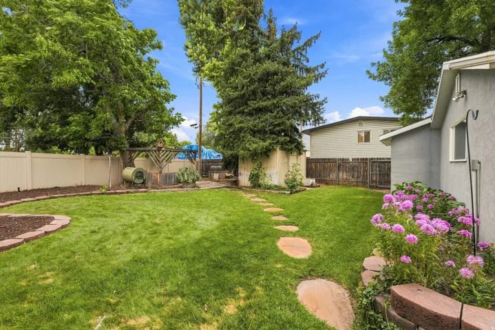 Expansive backyard featuring a lush lawn, mature trees, a stepping stone path, established garden beds with flowering plants, and a partial view of a detached structure