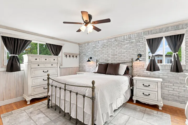 Bedroom featuring a whitewashed brick accent wall, wood-finish flooring, a ceiling fan with integrated lighting, and dual windows