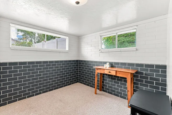 Carpeted room featuring a white ceiling, two horizontal windows with blinds, and white painted brick walls with a dark subway tile wainscoting