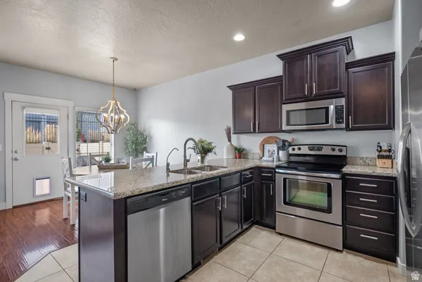 Open-concept kitchen featuring dark wood cabinetry, stainless steel appliances, a center island with sink, granite countertops, and light tile flooring