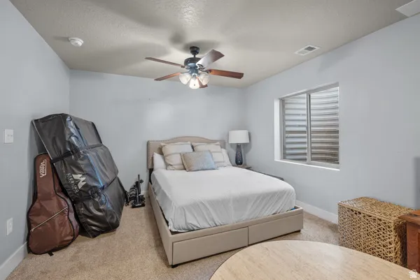 Carpeted bedroom featuring light gray walls, a window with a horizontal grate, and a ceiling fan with a wood-finish base and three lights