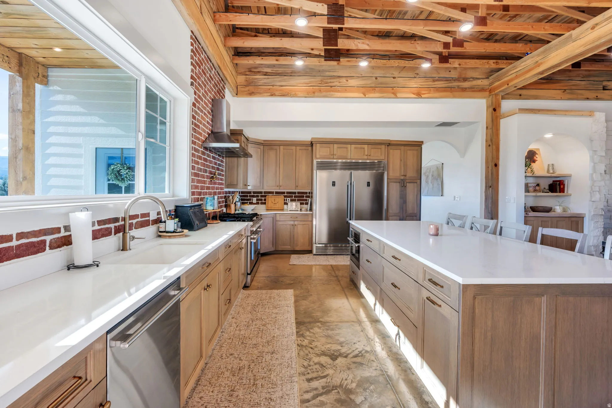 Spacious kitchen featuring an exposed wood beam ceiling, expansive kitchen island with a light-toned countertop, integrated stainless steel appliances, brick accent wall, and wood-finish cabinetry