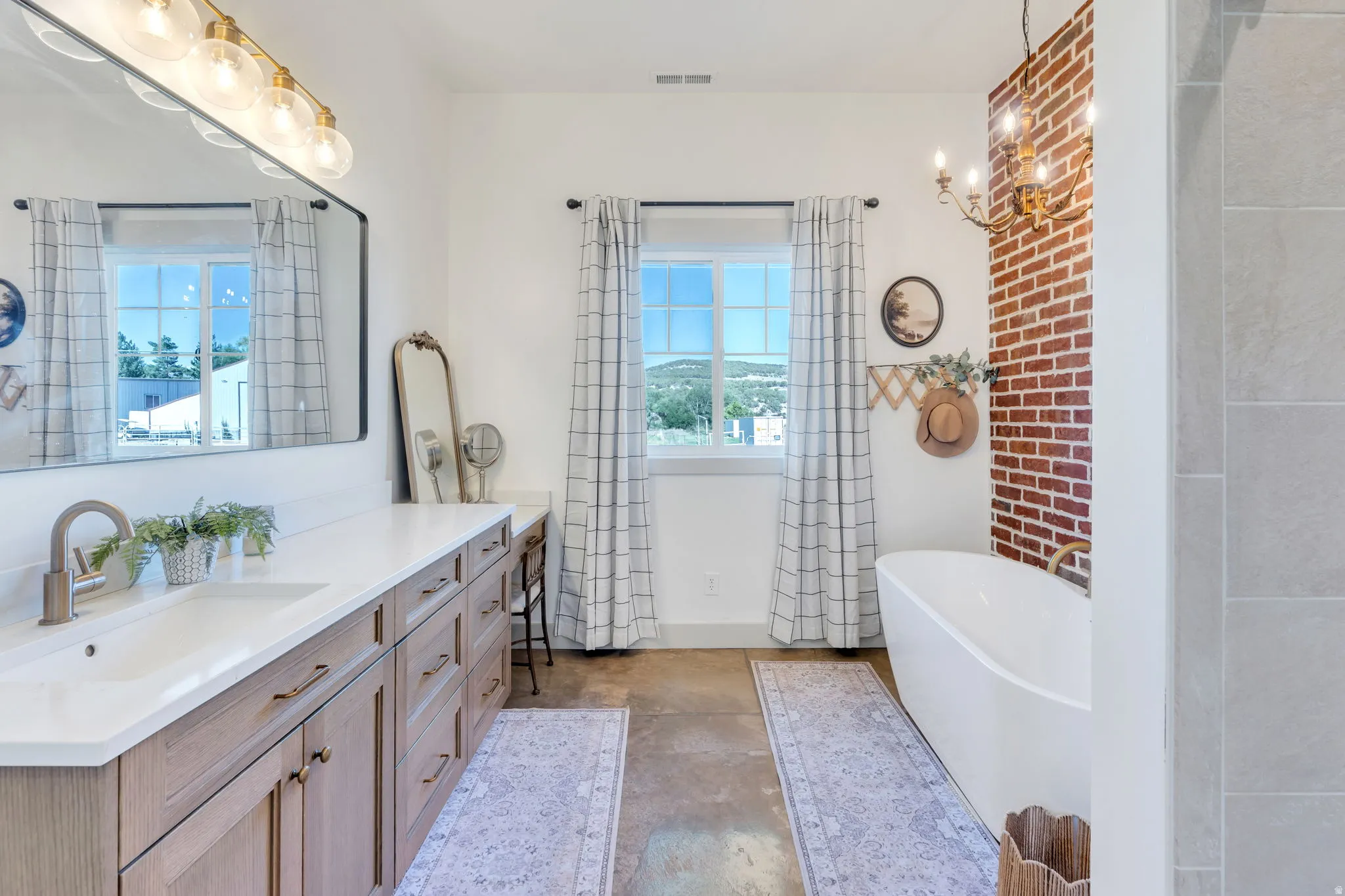 Spacious bathroom featuring a double vanity with wood-finish cabinetry and white countertops, a freestanding soaking tub, an exposed brick accent wall, and dual windows with natural light