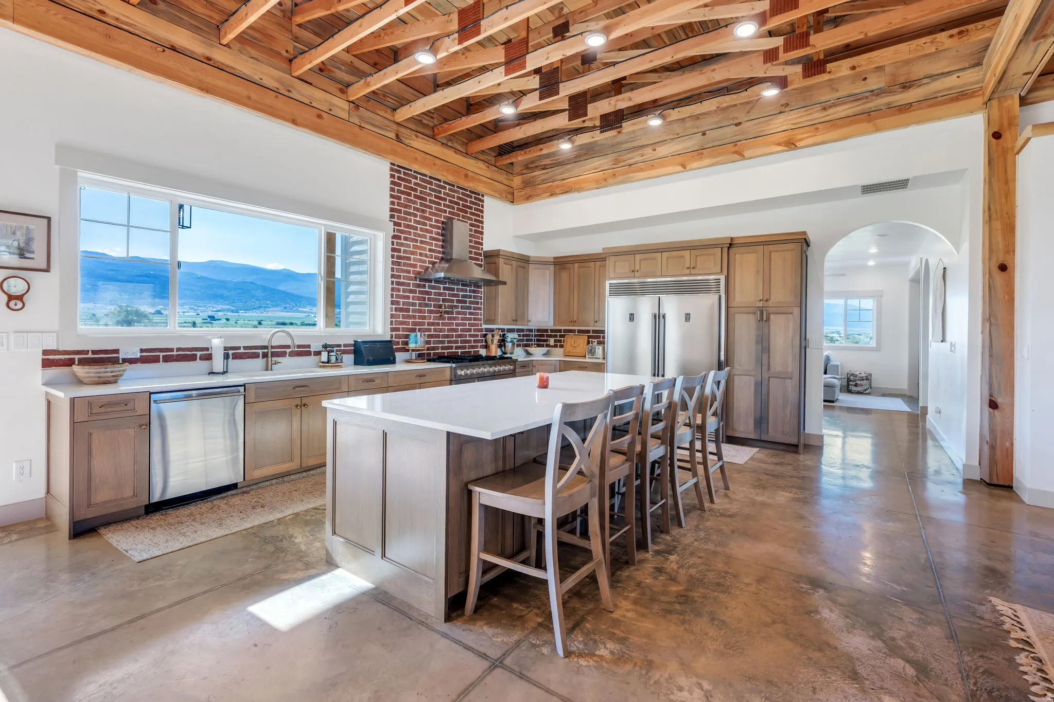 Spacious kitchen with exposed beam ceiling and recessed lighting, featuring an expansive center island with seating, ample cabinetry, stainless steel appliances, and a red brick accent wall