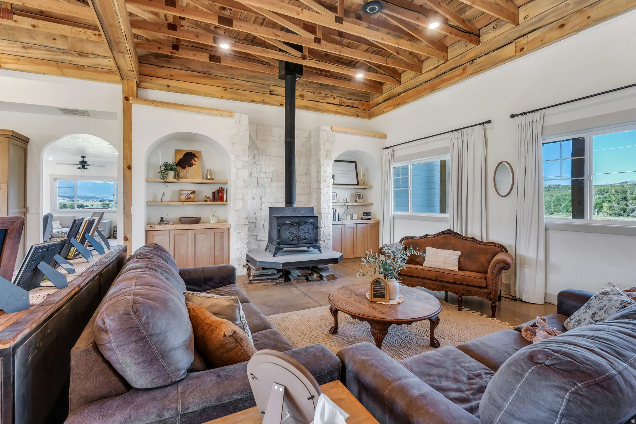 Vaulted wood beam ceiling with recessed lighting, stone-clad wood-burning stove, arched built-in shelving with base cabinetry, wood-finish flooring, and multiple windows