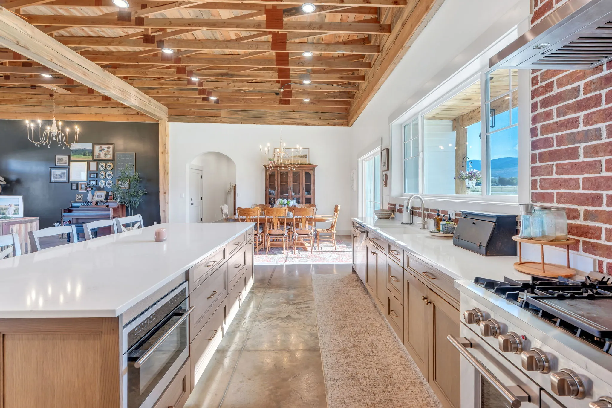 Open-concept kitchen with a large central island, exposed beam ceilings, industrial-style concrete flooring, and a brick accent wall with a stainless steel range and hood