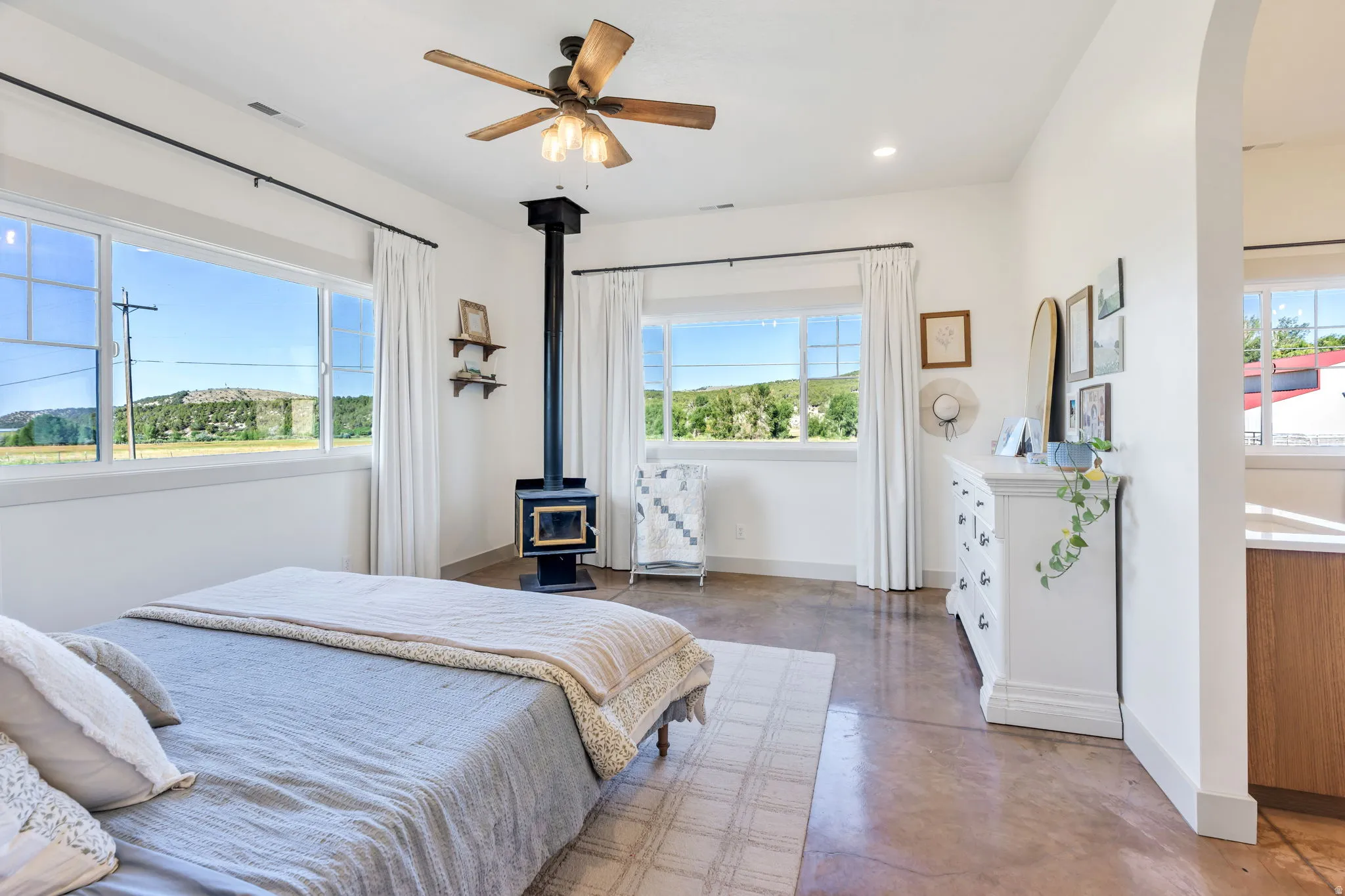 Spacious room featuring multiple windows, a ceiling fan with integrated lighting, a freestanding wood-burning stove, and polished concrete flooring