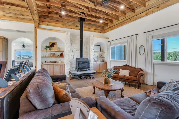 Vaulted wood beam ceiling with recessed lighting, stone-clad wood-burning stove, arched built-in shelving with base cabinetry, wood-finish flooring, and multiple windows