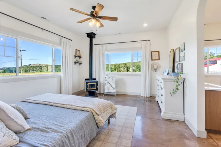 Spacious room featuring multiple windows, a ceiling fan with integrated lighting, a freestanding wood-burning stove, and polished concrete flooring