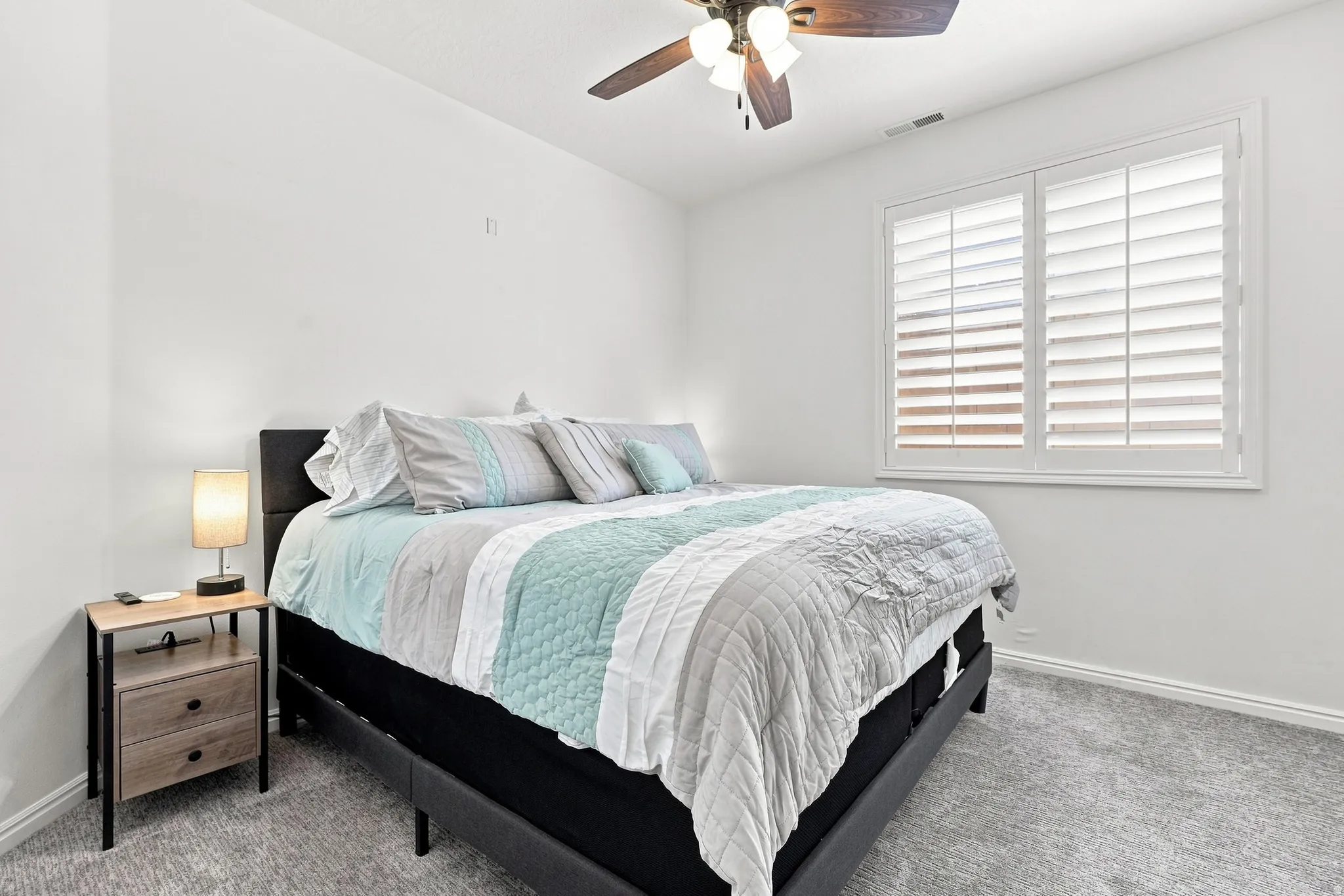 Carpeted room featuring a ceiling fan with light fixture, white walls, and a window with white plantation shutters