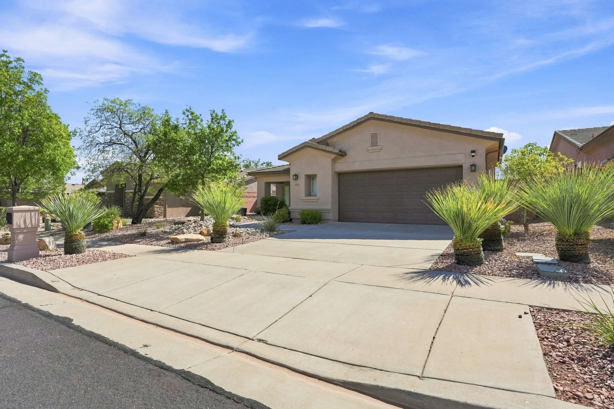 Desert landscape featuring mature trees and shrubbery, a concrete driveway, and a two-car garage