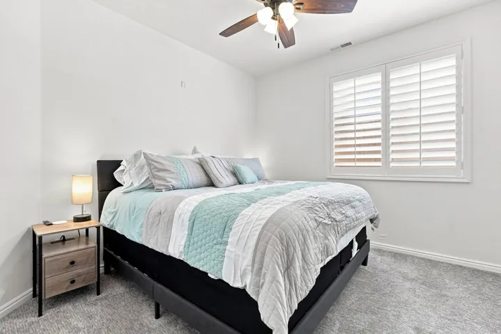 Carpeted room featuring a ceiling fan with light fixture, white walls, and a window with white plantation shutters
