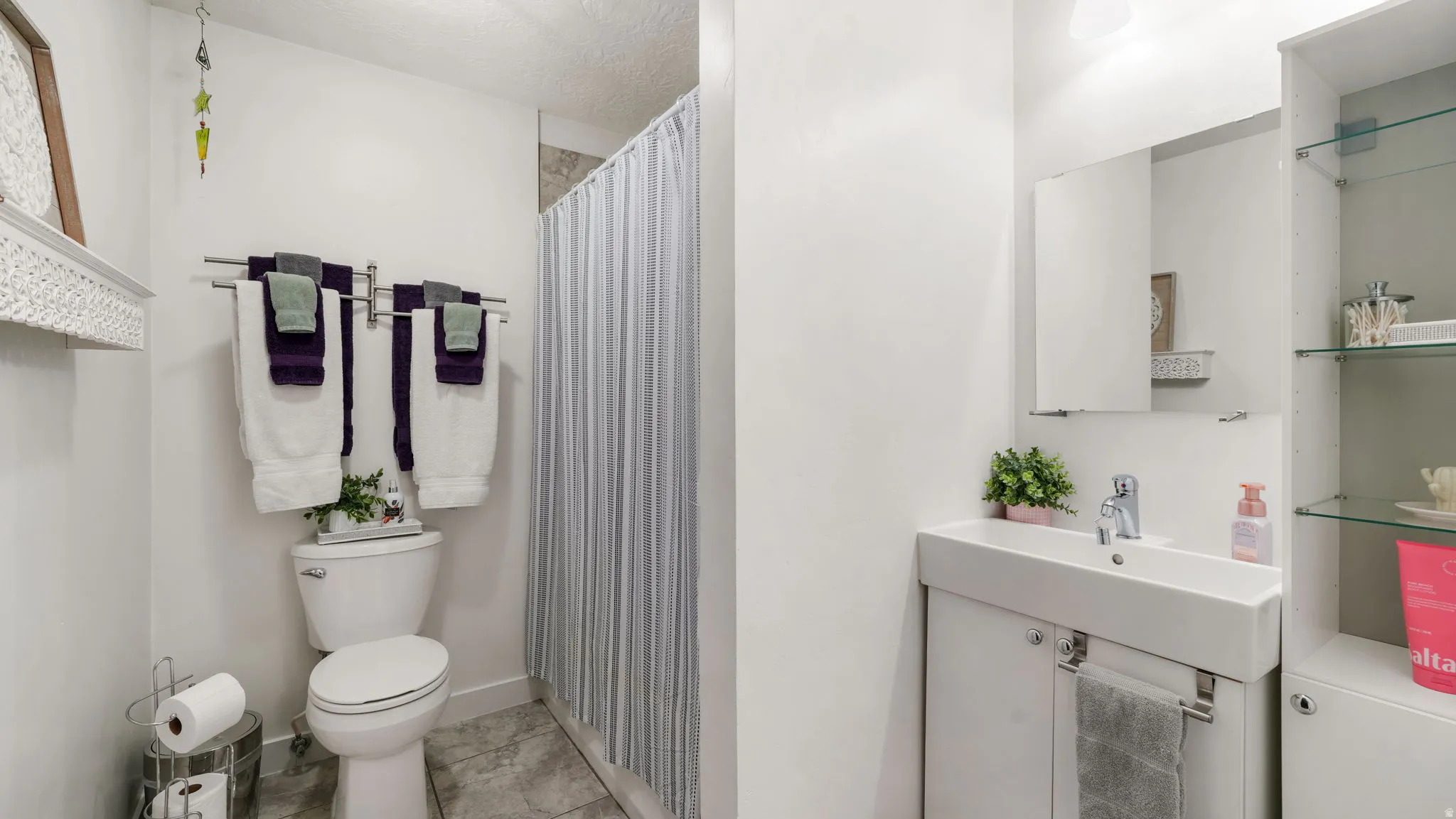Compact bathroom featuring a white toilet, a shower area with a striped curtain, a vanity with an integrated sink, a wall-mounted medicine cabinet, and a built-in storage unit with glass shelving