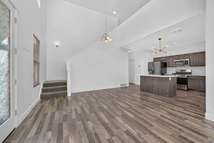 Open-concept living area featuring wood-finish flooring, a two-story ceiling, a staircase with carpeted steps, a modern kitchen island with a white countertop, and stainless steel appliances