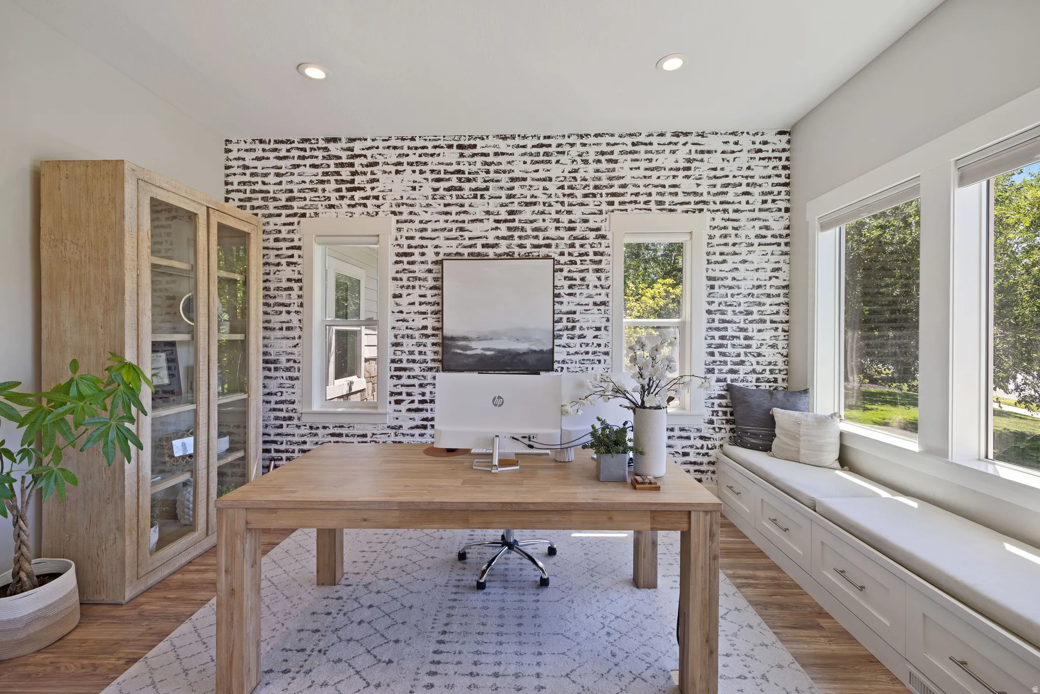 Office featuring LVP flooring, a prominent accent wall with a distressed brick pattern, and multiple windows and built in seating/storage