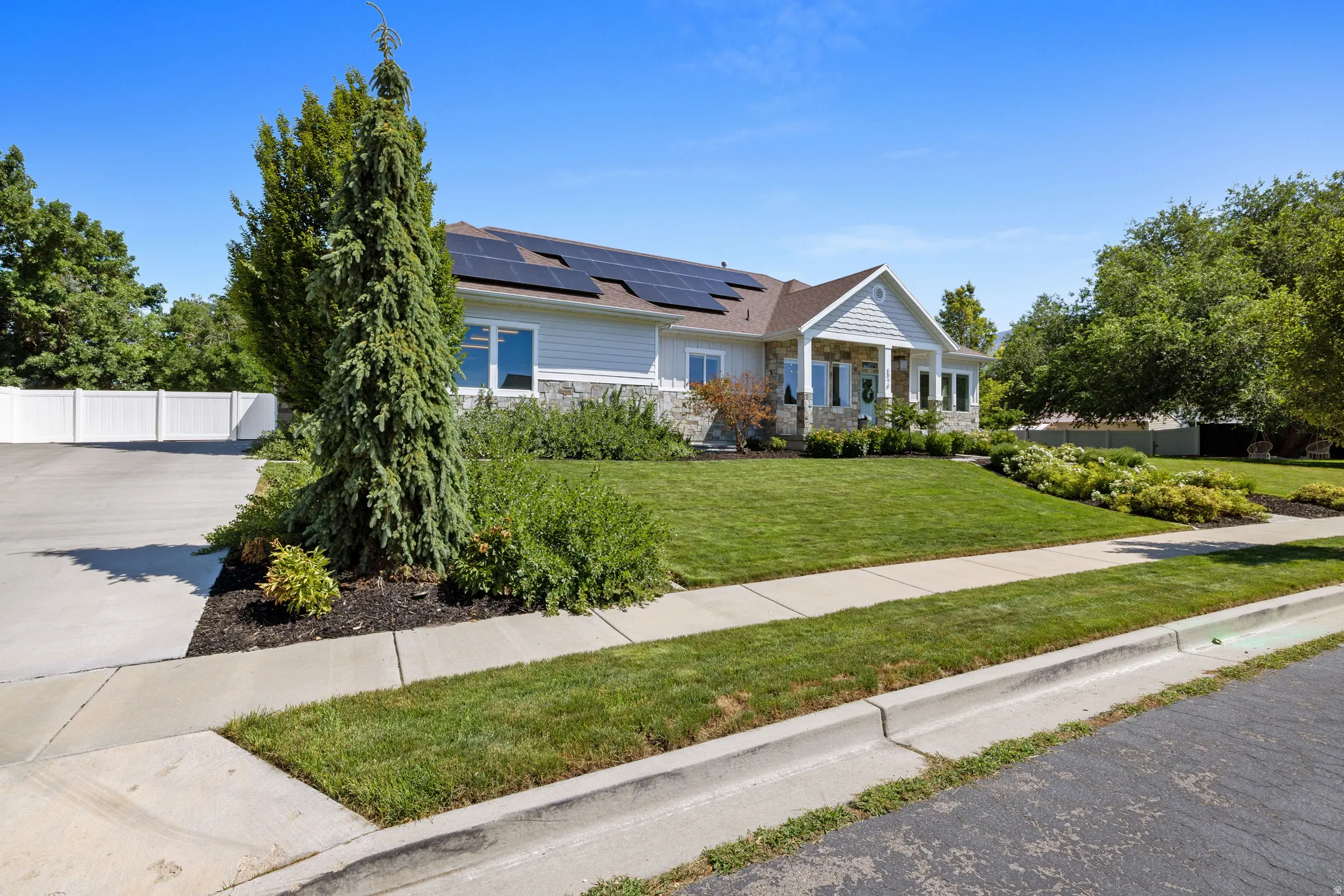 Ranch-style residence featuring a stone and siding exterior, solar panels, mature landscaping, a concrete driveway, and a white fence