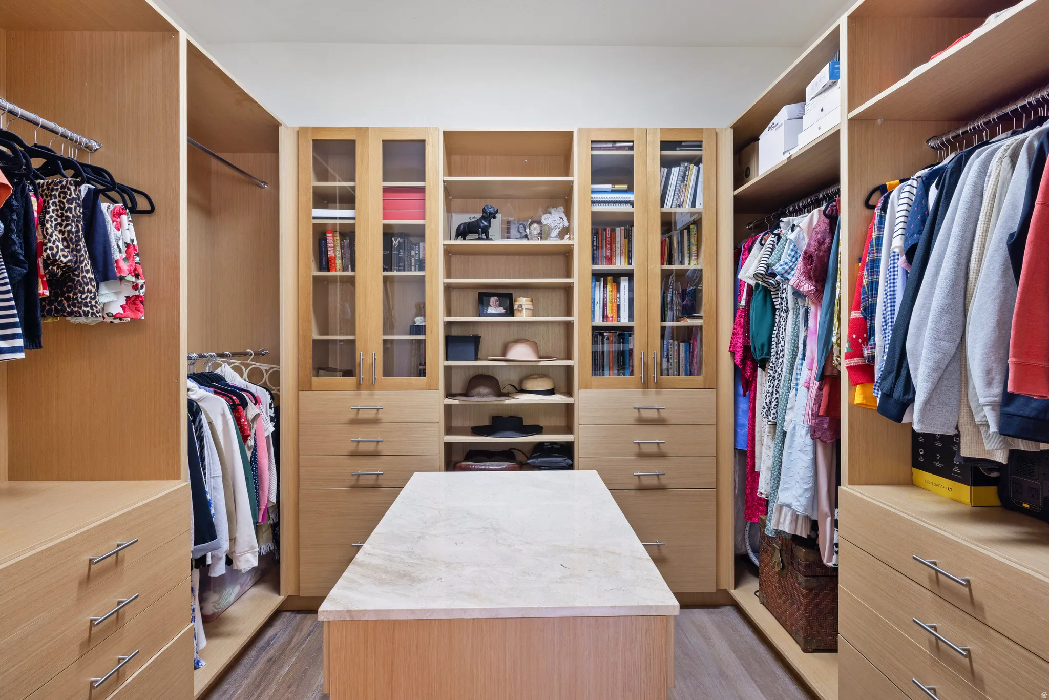 Primary walk-in closet featuring light wood-finish cabinetry, a central island with a stone-finish countertop, integrated shelving, hanging rods, and drawers with brushed nickel hardware