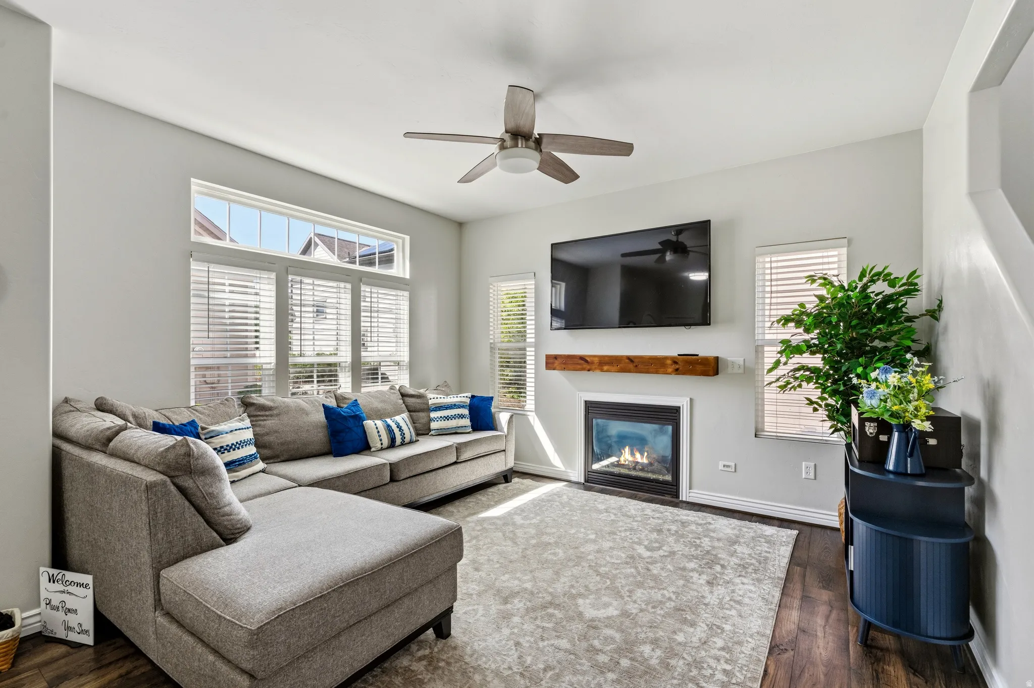 Gas fireplace with a wood-finish mantel, multiple windows with blinds, wood-finish flooring, and a ceiling fan with light fixture