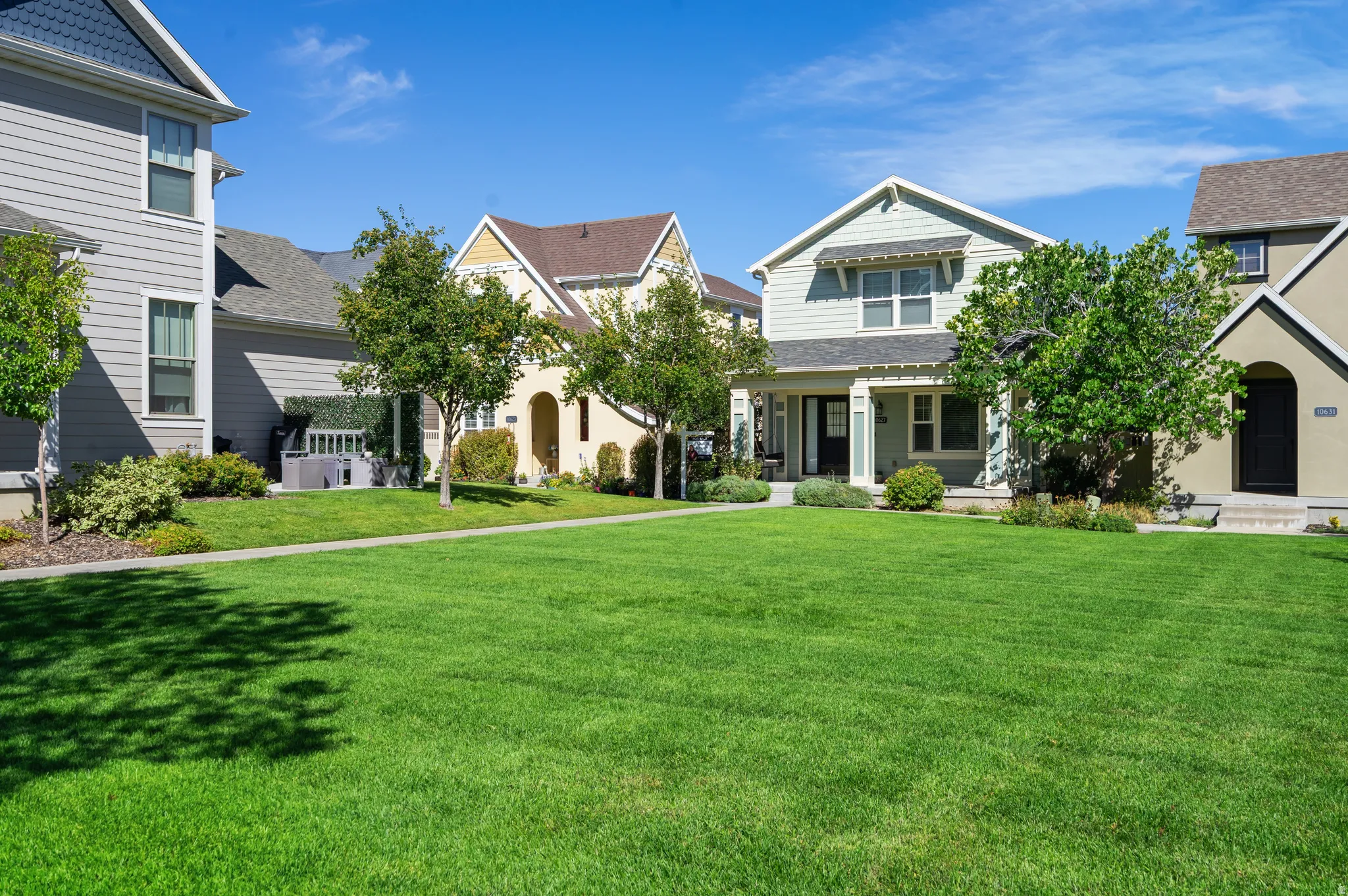 Expansive green lawn with mature trees and a sidewalk, bordered by a light blue home with a covered porch and a light yellow home with an arched entrance