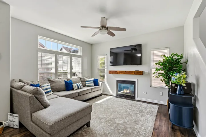 Gas fireplace with a wood-finish mantel, multiple windows with blinds, wood-finish flooring, and a ceiling fan with light fixture