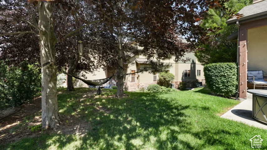 Manicured lawn area featuring mature shade trees, established shrubbery, a concrete patio, and a stucco exterior with stone accents