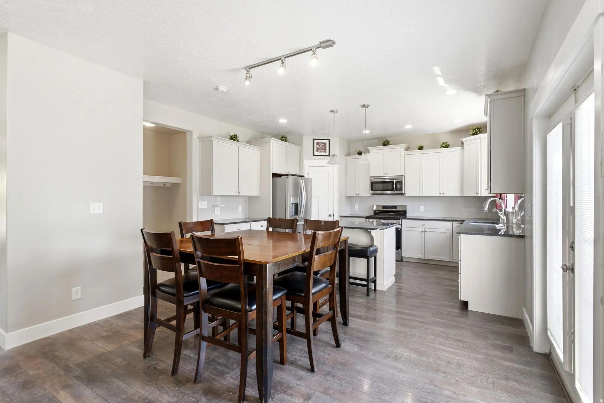 Spacious kitchen and dining area featuring wood-finish flooring, white cabinetry, dark countertops, stainless steel appliances, and track lighting