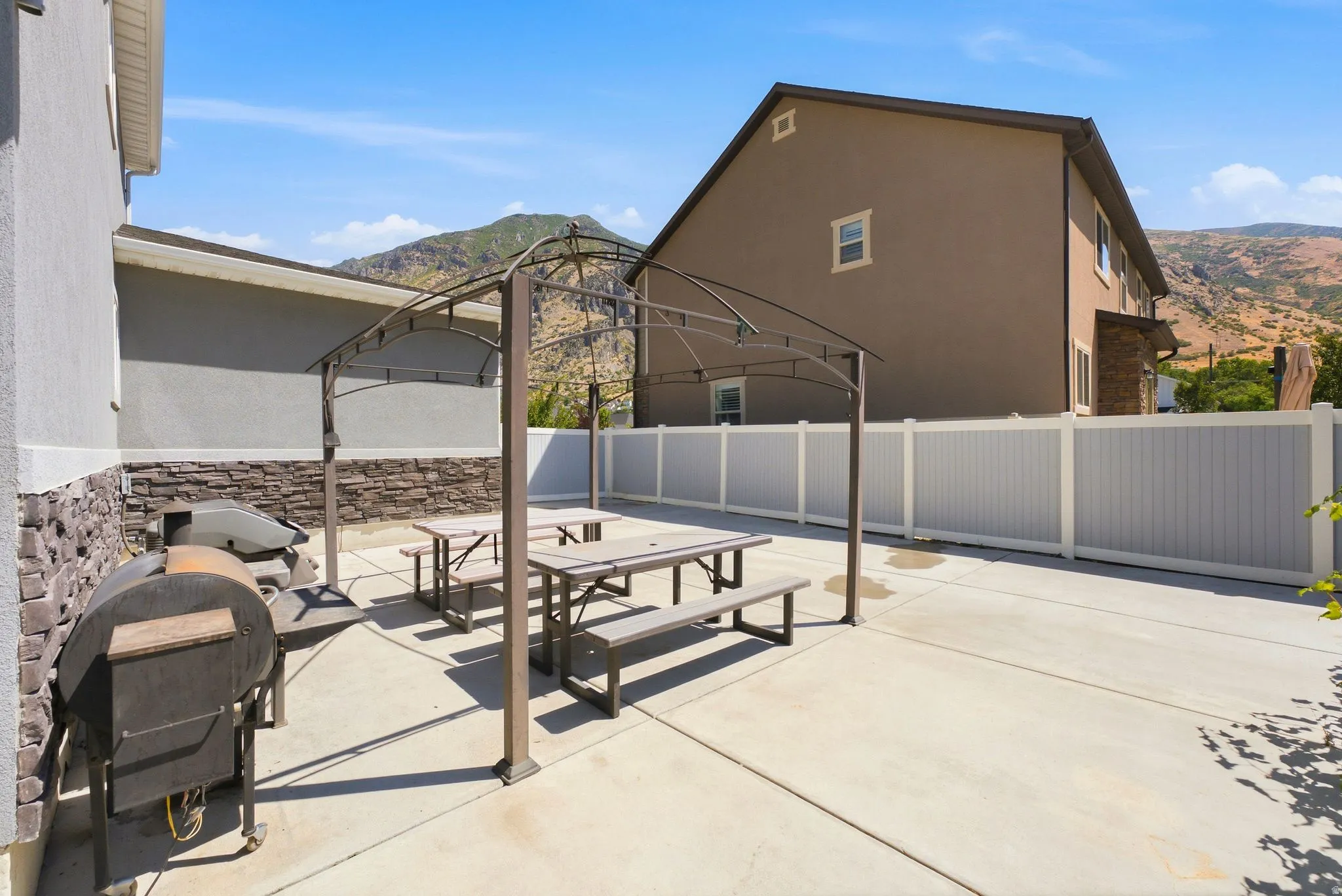 Expansive concrete patio area featuring a metal gazebo frame, two picnic tables with benches, and a privacy fence