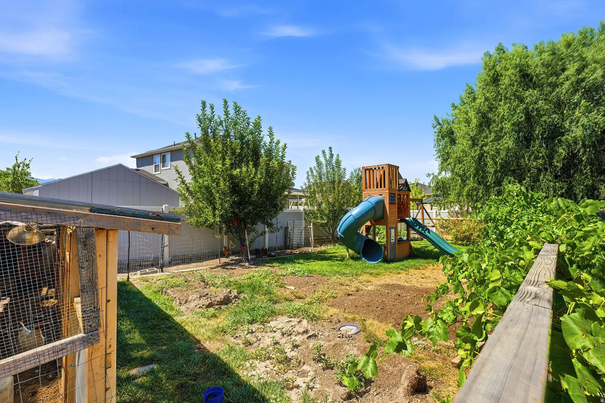 Expansive backyard space featuring mature trees, a wooden playset with a tube slide and open slide, and a wooden fence border