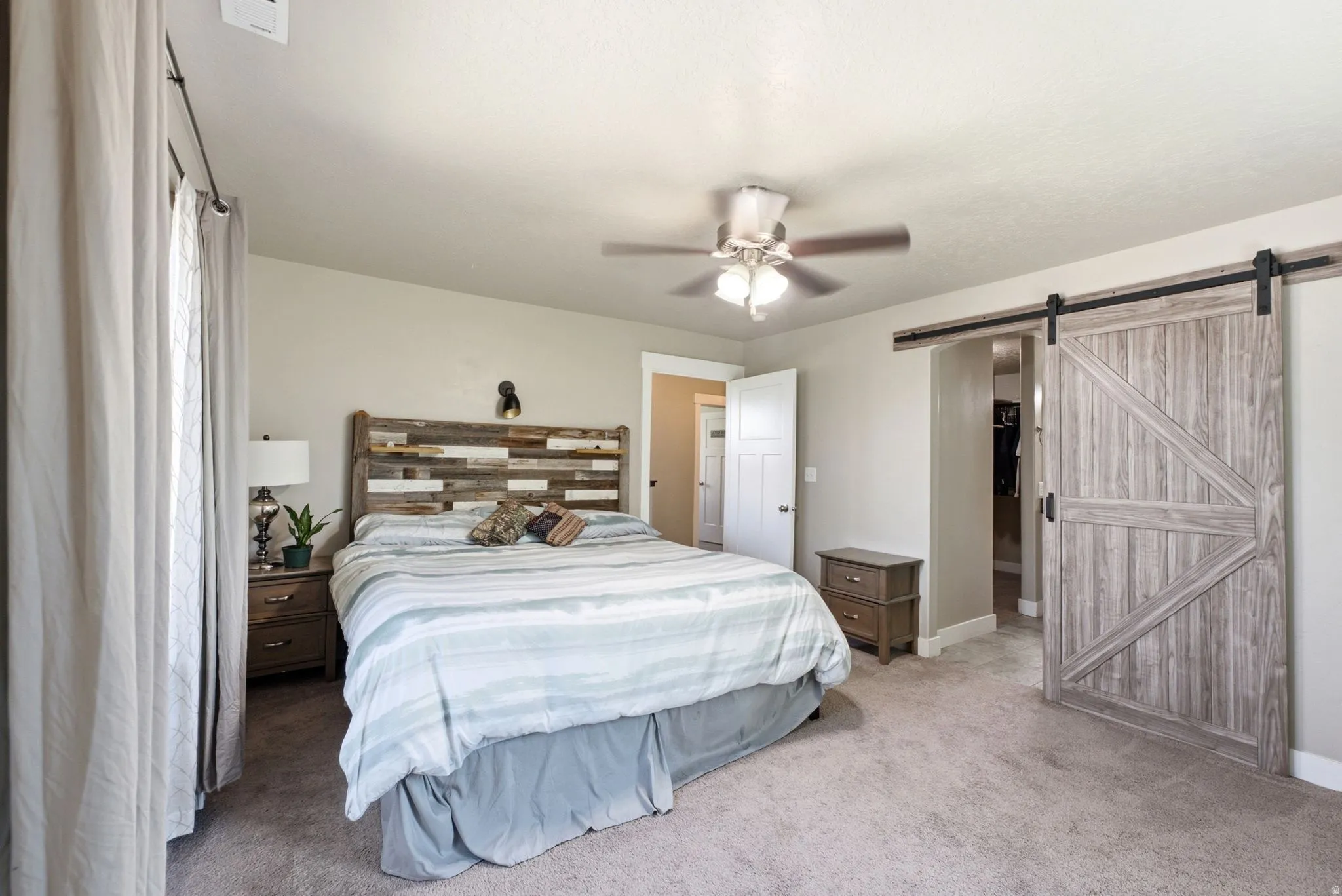 Expansive bedroom featuring a barn door entry, ceiling fan, carpeted flooring, and a large window with privacy curtains