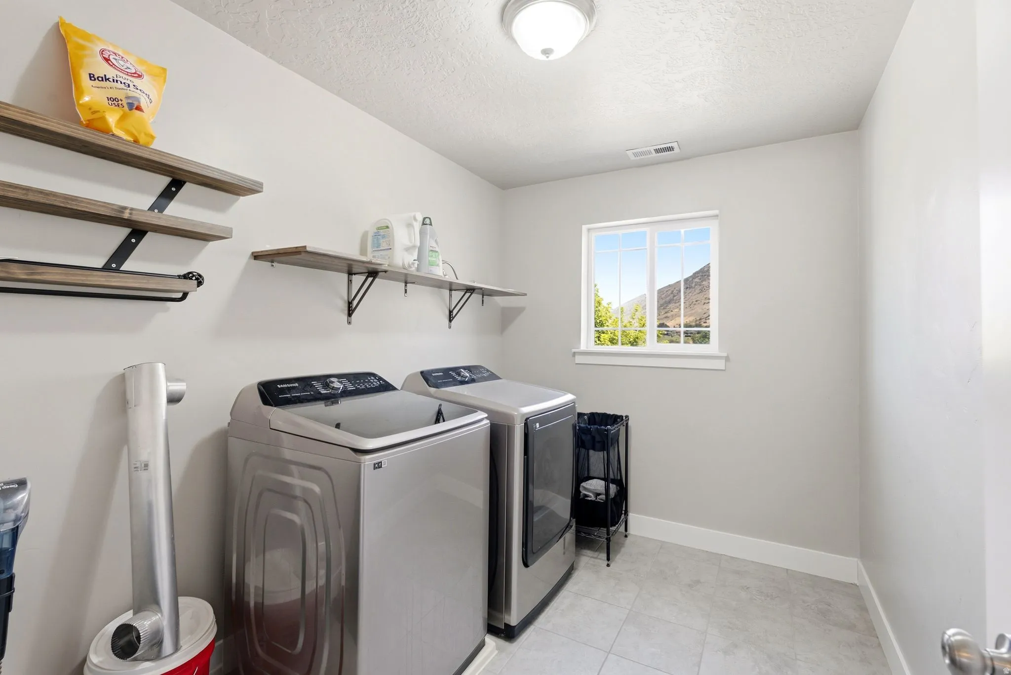 Laundry room featuring light gray walls, tile flooring, two wall-mounted wood-finish shelving units with black metal brackets, and a window with mountain scenery