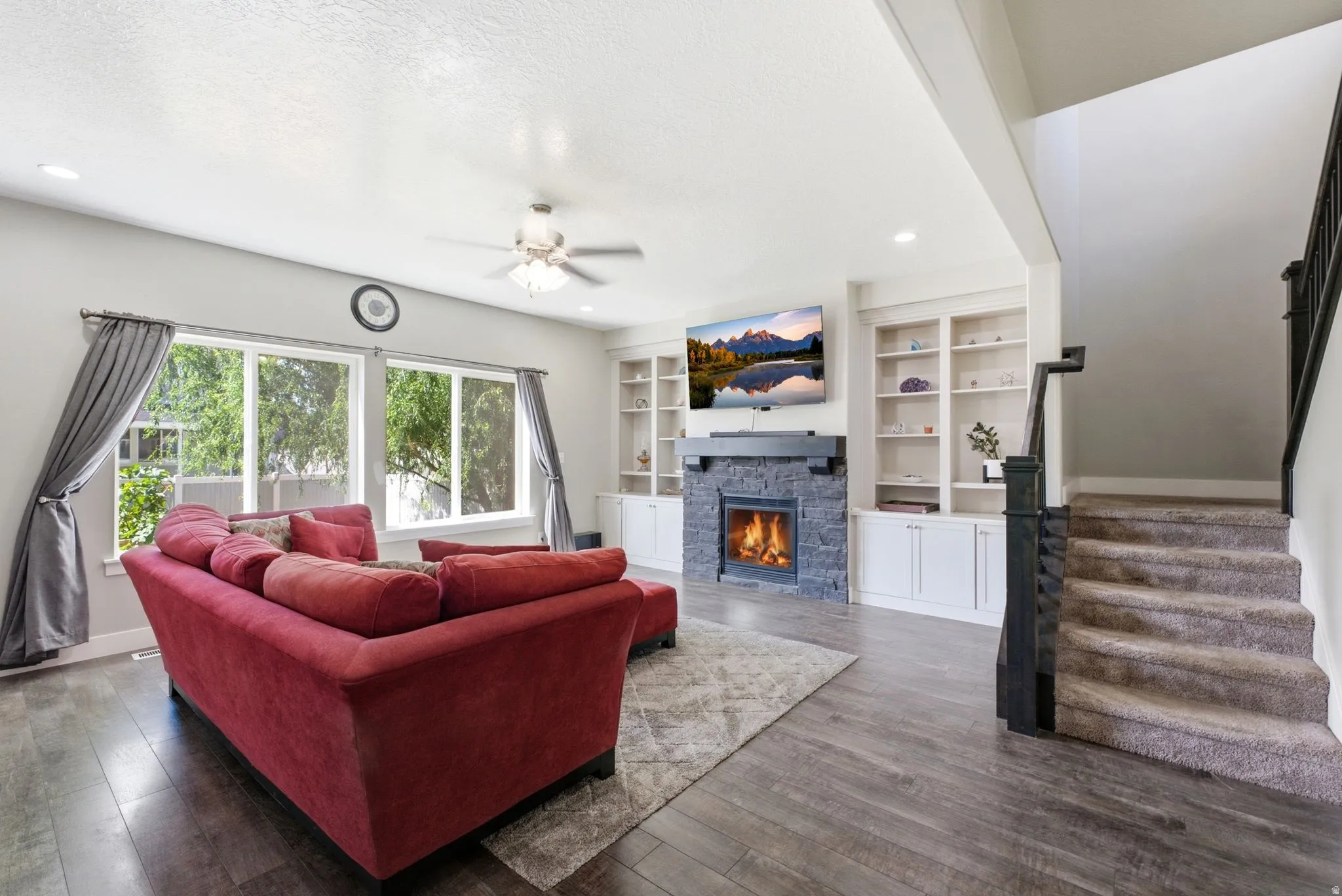 Living area featuring a stone-clad fireplace with mantel, built-in shelving, wood-finish flooring, a ceiling fan, and large windows