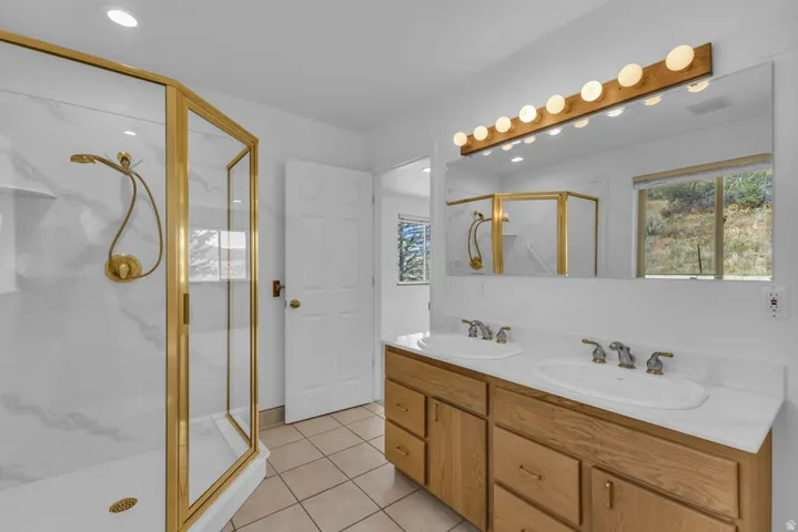 Contemporary bathroom featuring a corner glass shower enclosure with gold-toned framing and fixtures, a dual vanity with a light wood-finish cabinet, white countertops, and integrated sinks