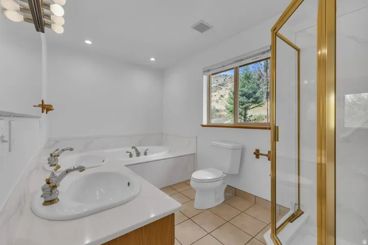 Double vanity with undermount sinks, a built-in jetted tub, a separate glass-enclosed shower with gold-tone fixtures, tile flooring, and a window with mountain scenery