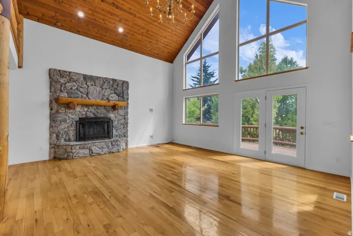 Vaulted wood-paneled ceiling, stone fireplace with log mantel, natural wood-finish flooring, and expansive windows with double French doors leading to an outdoor deck