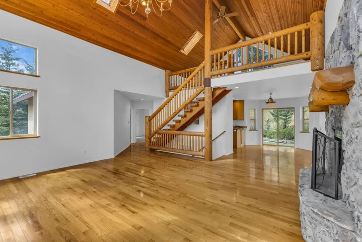Vaulted timber ceiling with exposed beams, wood-finish flooring, stone fireplace with log mantel, log-style staircase, and multiple windows