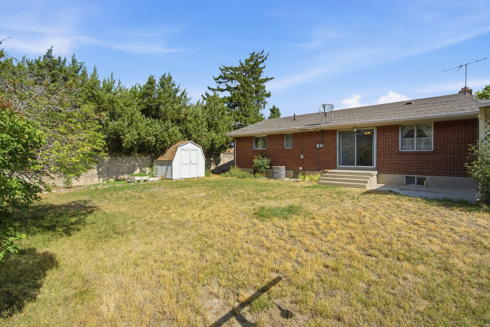 Expansive backyard featuring a brick exterior with sliding glass doors, concrete steps, a detached storage shed, and mature perimeter trees