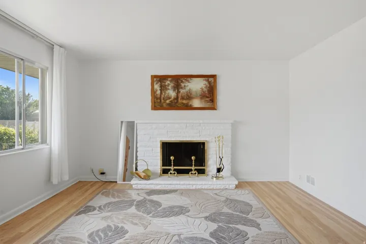 Living area featuring a painted brick fireplace with a brass firebox, light wood-finish flooring, white baseboards, and a large sliding window