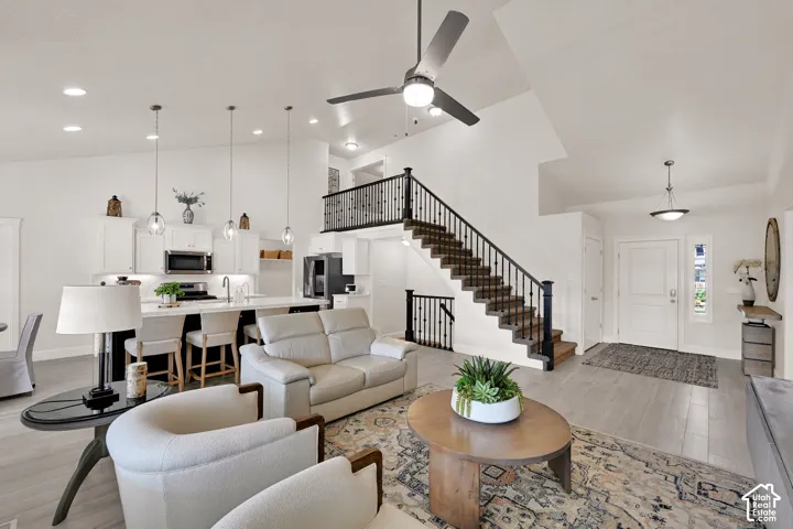 Bright open-concept living space featuring high ceilings, light wood-finish flooring, a modern kitchen with a large island, a staircase with dark wood treads and iron balusters, and a contemporary ceiling fan