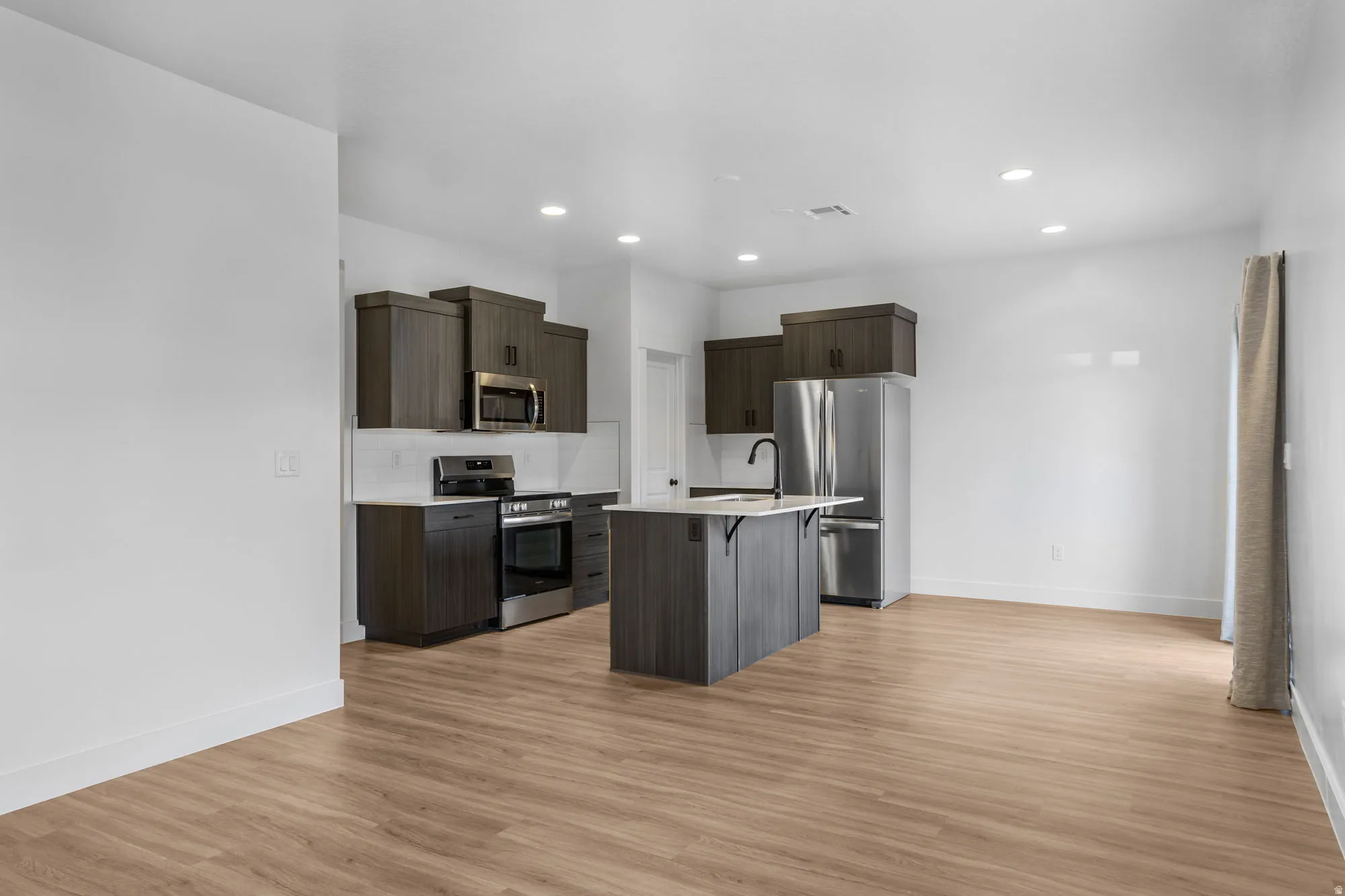 Open-concept kitchen featuring dark wood-finish cabinetry, stainless steel appliances, a center island with a sink, white countertops, and wood-finish flooring