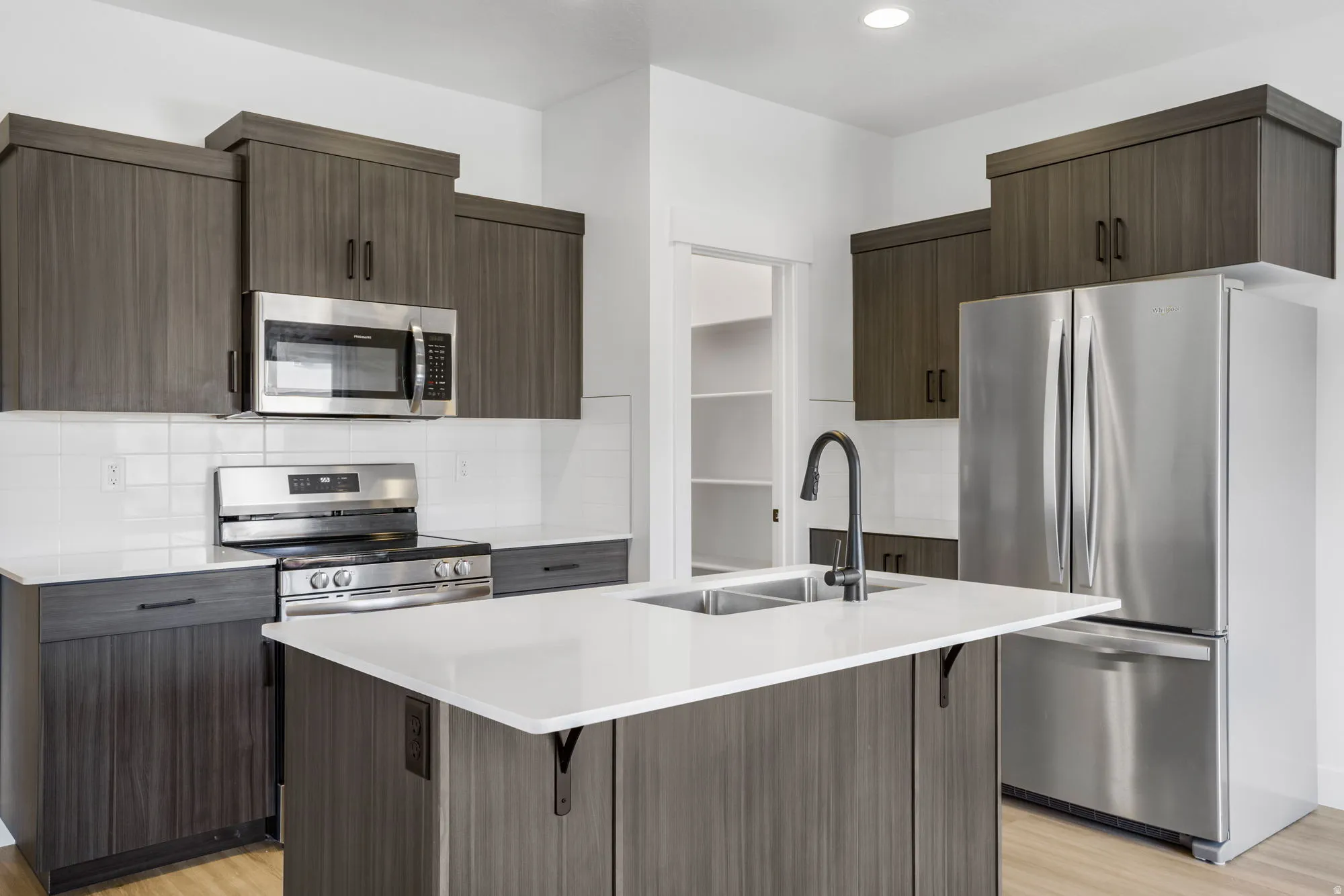 Modern kitchen featuring dark wood-finish cabinetry, stainless steel appliances, white subway tile backsplash, a center island with double sink, and light-toned flooring