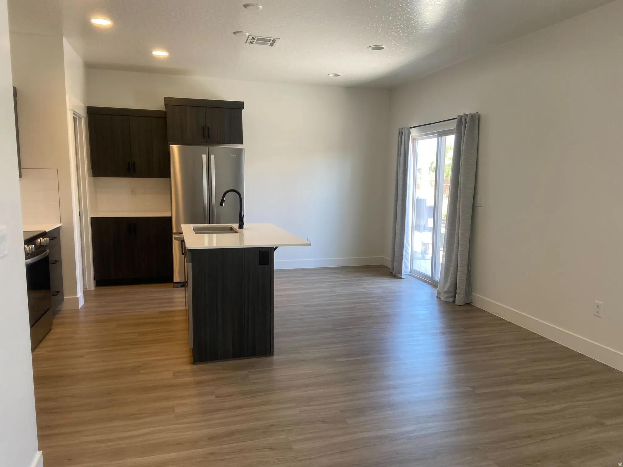 Modern kitchen and living area featuring dark wood-finish cabinetry, stainless steel appliances, a center island with a white countertop and integrated sink, wood-finish flooring, and a sliding glass door providing exterior access