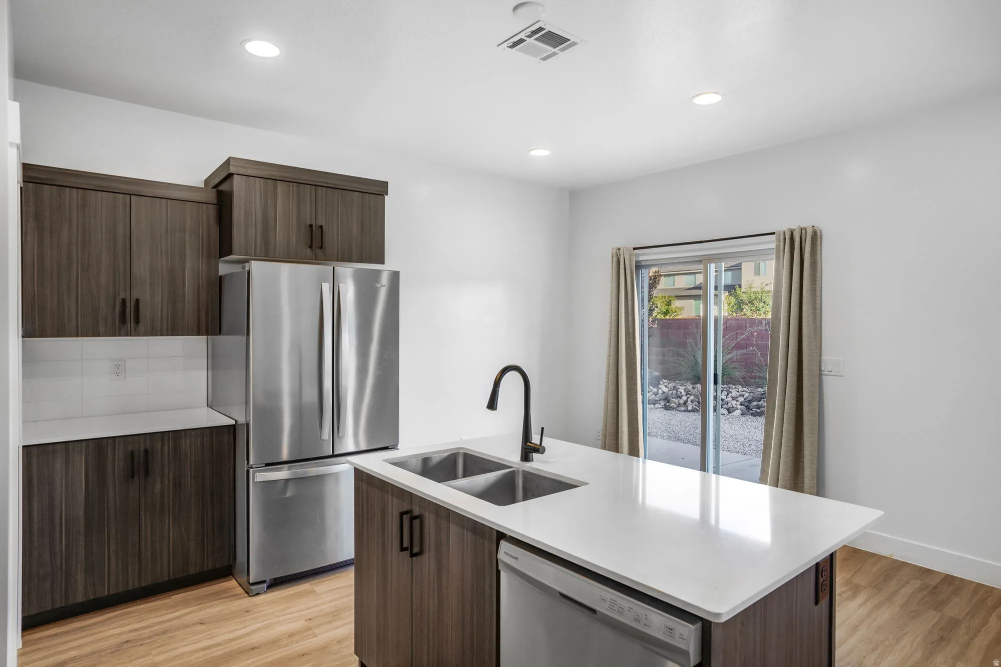 Modern kitchen featuring an island with a double basin sink, white countertop, dark wood-finish cabinetry, stainless steel appliances, and wood-finish flooring