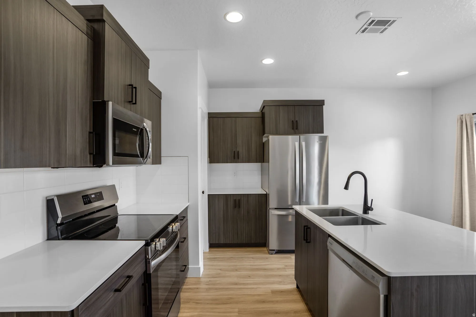 Modern kitchen featuring wood-finish cabinetry, stainless steel appliances, a white tile backsplash, light countertops, and wood-finish flooring