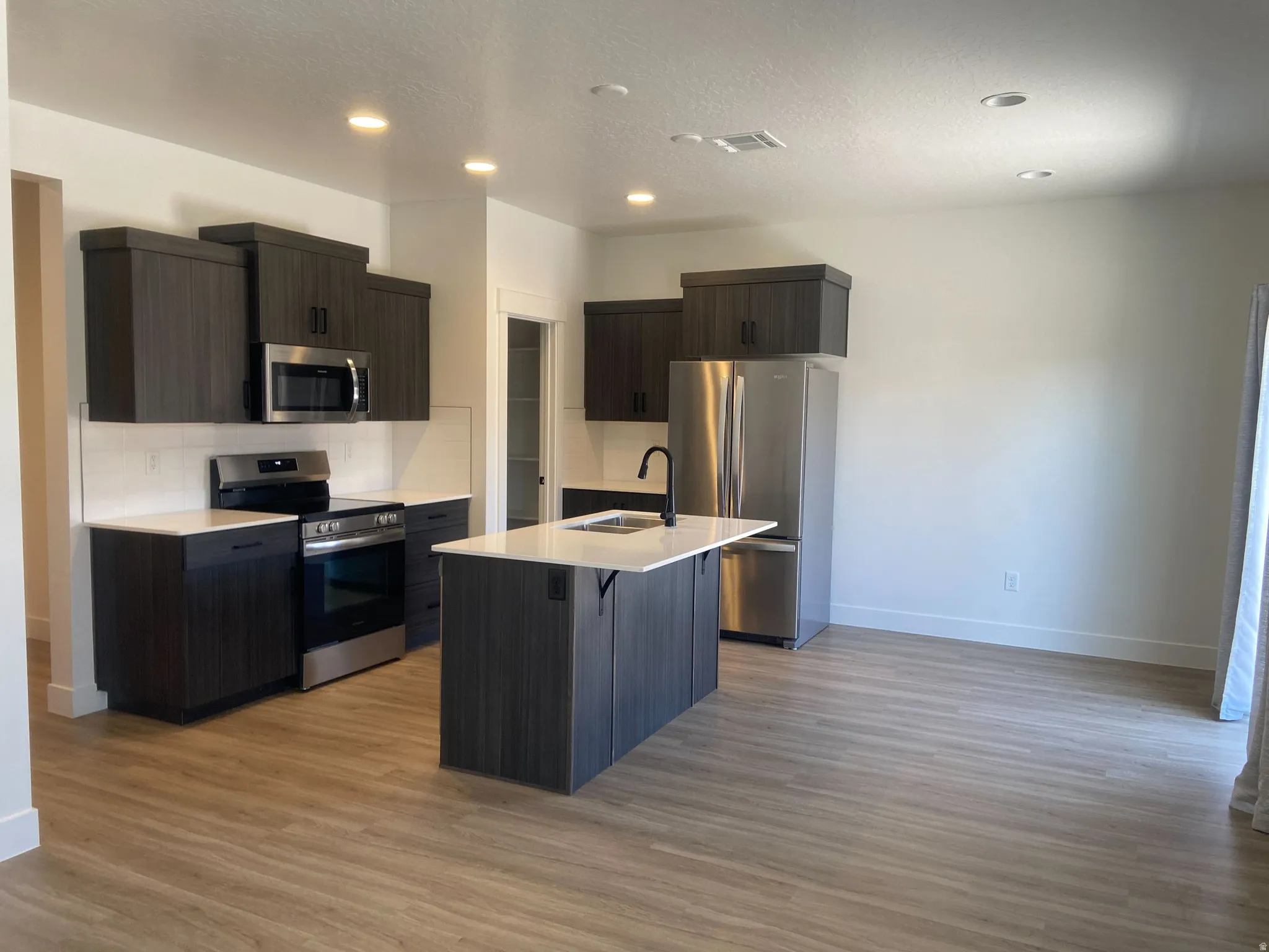 Contemporary kitchen featuring dark wood-finish cabinetry with stainless steel appliances, a center island with a sink, white countertops, and wood-finish flooring